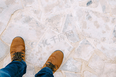 踩摄影照片_男性脚穿着棕色靴子和蓝色牛仔裤，踩在浅色石砖上。