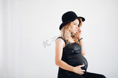 戴着黑帽子和闪闪发光的上衣拥抱她的腹部的时尚孕妇。