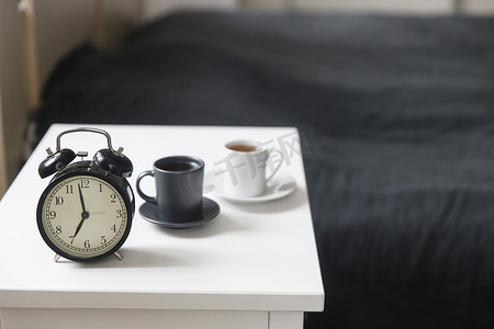 图片闹钟，床前的白色床头柜上放着一杯黑咖啡，床前配有灰色床单。