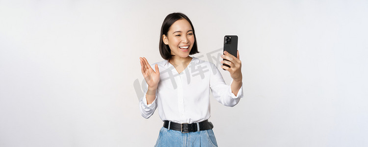 时尚的现代亚洲女性在智能手机前置摄像头挥手、视频聊天、在手机应用程序上与人聊天、白色背景的形象