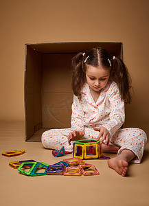 穿着睡衣、扎着两个马尾辫的欧洲小女孩玩着米色背景的彩色磁性构造玩具，上面有复制广告空间