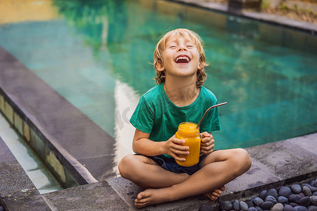 水池背景摄影照片_男孩在玻璃梅森罐子里喝着多汁的芒果冰沙，水池背景上有钢吸管。