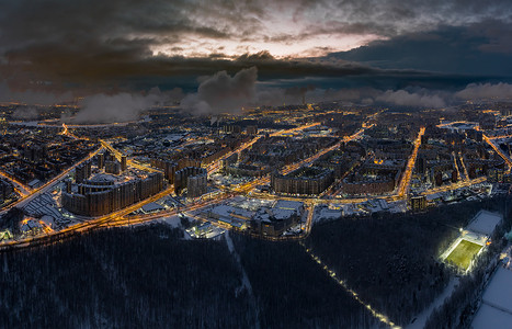 公园与建筑摄影照片_圣彼得堡-俄罗斯黄昏时的空中冬季城市景观，无人机飞过巨大的住宅区和公园，建筑起重机，夜间照明，云彩漂浮在无人机下方