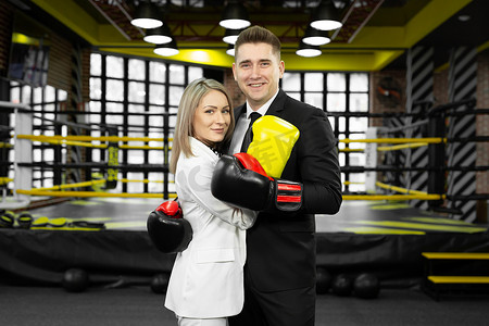 拳击场摄影照片_年轻的已婚夫妇，一男一女穿着西装，戴着拳击手套，在拳击场的对面拥抱。