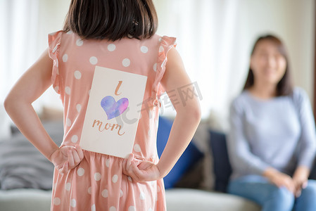 亚洲可爱的女孩隐藏手工制作的贺卡，上面写着“我爱妈妈”的话，让她在家给妈妈带来惊喜