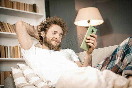 躺沙发摄影照片_卷发男子躺在沙发上，看着手机屏幕，查看社交媒体。