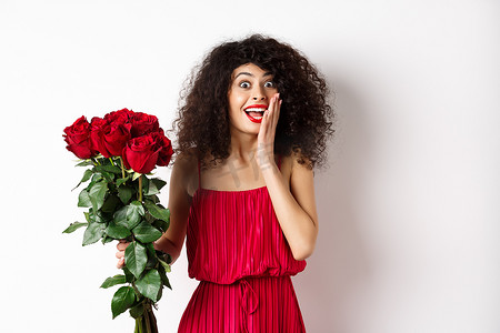 穿着红裙的兴奋的卷发女人，收到玫瑰花束，看上去很惊讶，为浪漫的礼物而高兴，站在白色背景上