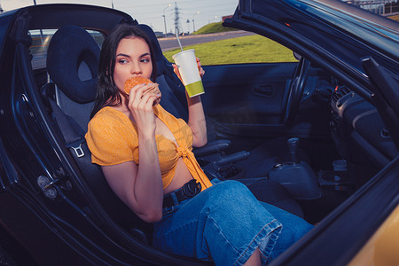 穿着蓝色牛仔裤和橙色上衣的模特坐在黄色汽车敞篷车里吃汉堡，拿着纸杯里的饮料。