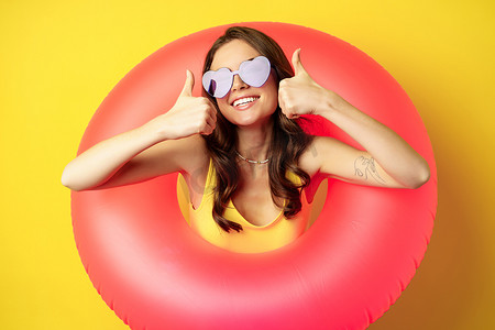 游泳圈的女孩摄影照片_戴墨镜、戴着粉色游泳圈、微笑、竖起大拇指、站在黄色背景上的快乐迷人女性的特写