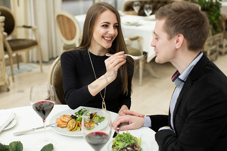 互相喂食摄影照片_美丽的年轻夫妇在餐厅里一边互相喂食，一边微笑。