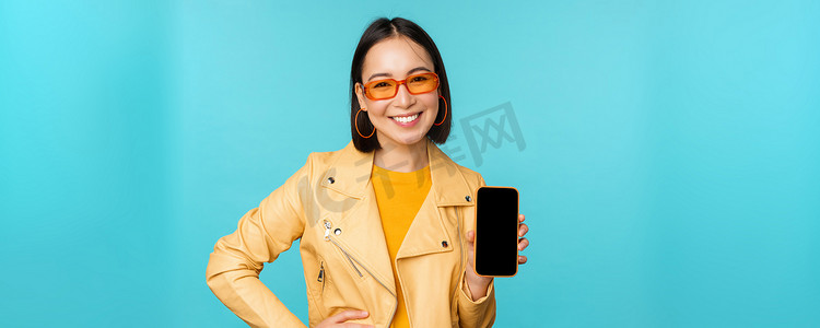 年轻时尚的亚洲女模特戴着时尚太阳镜的形象，显示手机屏幕，智能手机界面上的应用程序，站在蓝色背景上