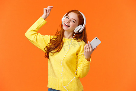 无忧无虑的快乐红发少女收到新耳机作为圣诞礼物，一边跳舞一边举手听最喜欢的音乐，拿着智能手机，放上很棒的曲目，橙色背景