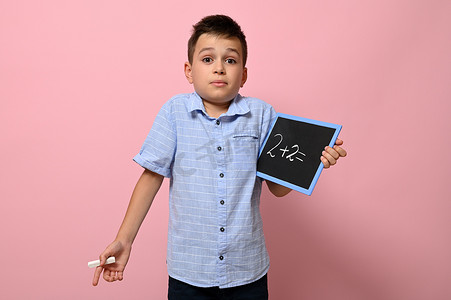 儿童数学摄影照片_一个可爱的男生手里拿着一块黑板，上面有算术问题，站在粉红色的背景上，有文字空间，带着惊讶和疑问看着镜头。