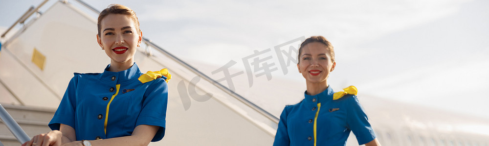 两位身穿蓝色制服的漂亮空姐对着镜头微笑，站在楼梯上欢迎乘客