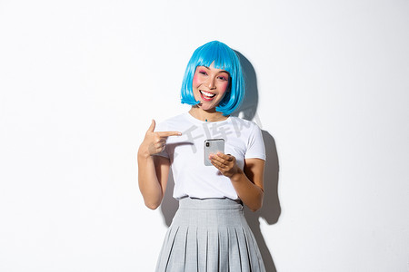 美丽的亚洲女孩的肖像，打扮成动漫人物，戴着蓝色短假发，用手指指着智能手机，微笑着，站在白色背景上
