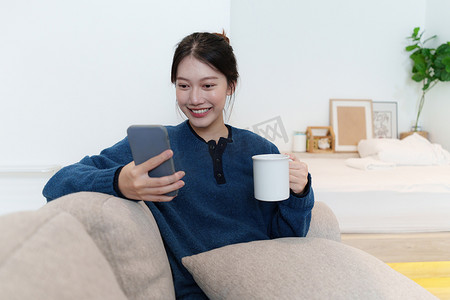 年轻的亚洲女性坐在沙发上与朋友聊天并通过智能手机查看社交媒体的肖像。