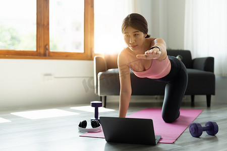 在家中穿着运动服的健康运动亚洲女性使用笔记本电脑在社交媒体上分享锻炼结果，并在健身垫上进行腹肌练习