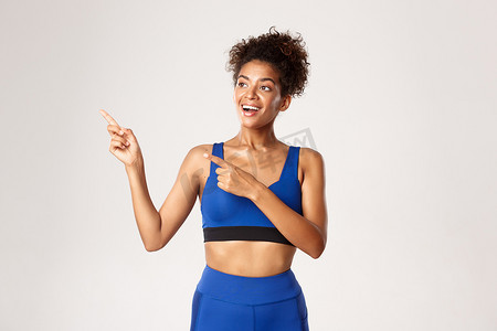 工作室拍摄的快乐健康的非洲裔美国健身女孩，身穿蓝色运动服，手指向左，面带高兴的微笑，站在白色背景下