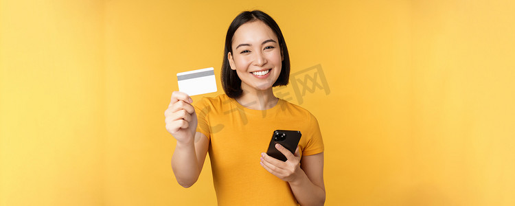 快乐的亚洲女孩微笑着，展示信用卡和智能手机，推荐手机银行，站在黄色背景下