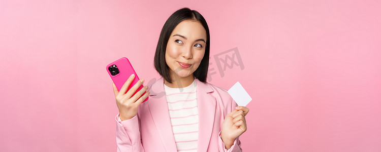 女士西装摄影照片_微笑的亚洲企业女性，穿着西装思考的女士，拿着智能手机和信用卡，计划在网上购买东西，用手机购物，粉红色背景