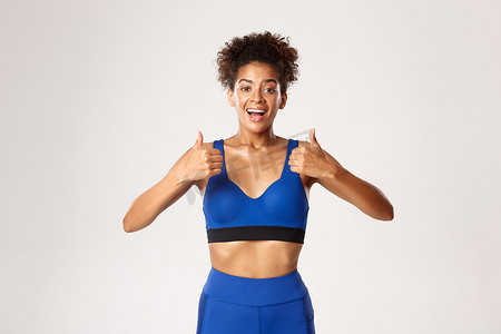 蓝色运动服摄影照片_身穿蓝色运动服、快乐健康的女性腰部以上，看起来很满意，竖起大拇指表示赞同一些好的白色背景