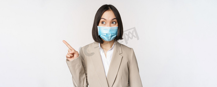 热情的女商人手指向左，戴着 covid-19 大流行的医用口罩，站在白色背景上