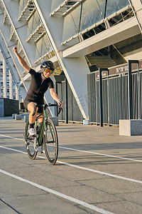 行服摄影照片_身穿黑色骑行服和防护装备的快乐职业女自行车手微笑着举起手臂，看起来对阳光明媚的户外训练结果感到满意