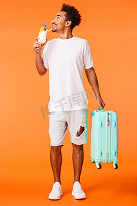 全长垂直拍摄梦幻般快乐轻松的非洲裔美国男子携带行李、手提箱和喝鸡尾酒，高兴地环顾四周，微笑着，在豪华酒店预订，橙色背景