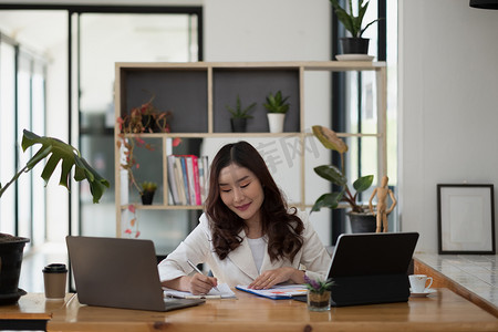 年轻的亚洲女性坐在办公桌前，拿着笔记本电脑做笔记的肖像。
