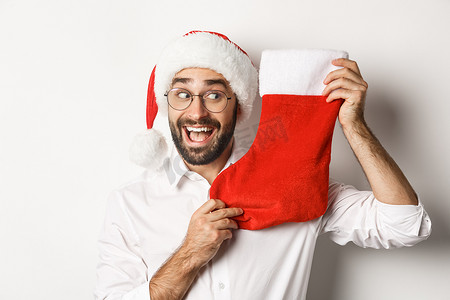 圣诞袜里有礼物摄影照片_快乐男人庆祝圣诞节的特写，收到圣诞袜里的礼物，看上去很兴奋，戴着圣诞帽和眼镜，白色背景