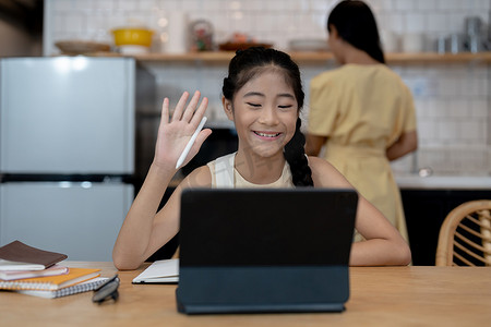 戴耳机的微笑亚洲小女孩使用笔记本电脑与老师进行远程视频通话，快乐的小孩与导师挥手致意，在电脑上在线学习，在家上学的概念