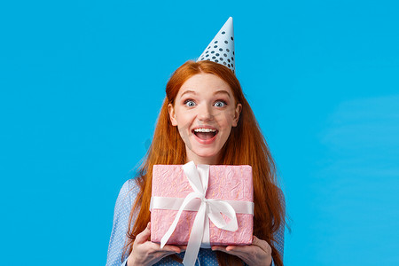 兴奋漂亮的女性、迷人的红发女人，蓝眼睛快乐的表情，收到可爱的粉红色礼物，礼物包裹在盒子里，微笑着逗乐，戴着生日帽庆祝生日
