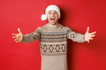 穿着圣诞毛衣和圣诞帽的快乐、有魅力的男人的肖像，伸手去拿礼物，想要拿点东西，微笑着，站在红色背景上
