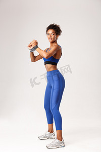 身着蓝色运动服、用橡皮筋做运动、看着别处、在白色背景下锻炼的迷人非洲裔美国健身女孩的全长侧面照片