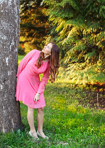 穿着粉红色裙子的年轻女子，靠在树上，抬起头来，背景是日落灯光公园。