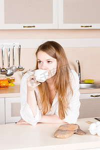 女孩穿着白色男式衬衫，长发飘逸，正在厨房喝茶，手肘放在桌子上。