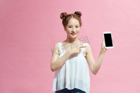 穿着夏季服装的快乐小女孩用手指指着粉红色背景中突显的空白屏幕手机