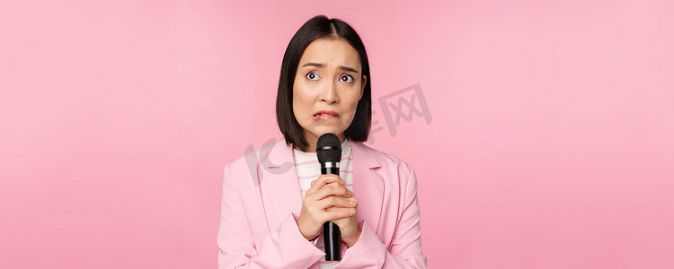 身穿西装、焦虑的亚洲女士在公共场合讲话，在会议上用麦克风发表演讲，看上去很害怕，站在粉红色的背景下