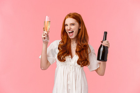 兴奋而无忧无虑的喝醉的红发白人女友玩得很开心，在很棒的派对上狂野而自由，举起玻璃杯和瓶子香槟，微笑着时髦，站在粉红色的背景上