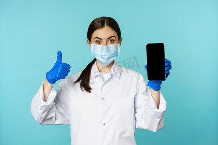 戴着医用面罩和手套的医生肖像，显示手机应用程序、智能手机屏幕和拇指，推荐在线检查网站，站在蓝色背景上