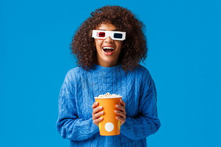 开朗华丽的非洲裔美国女性喜欢在电影院看电影，对 3D 效果兴奋地笑着，戴着眼镜，拿着爆米花，站在蓝色背景上