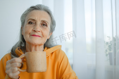 金发带摄影照片_快乐活泼的中年金发女人拿着一杯茶或咖啡，看着镜头，脸上带着灿烂的温暖微笑