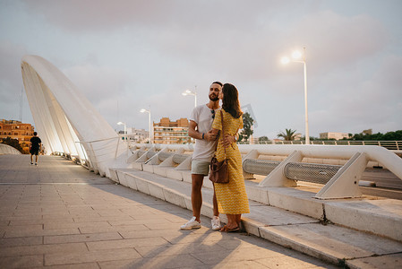 一个穿黄色裙子的女孩和她微笑的男朋友拥抱在一座白色的桥上