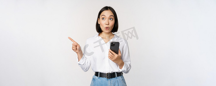 兴奋的亚洲女孩在移动应用程序上购物，拿着手机，用手指指着空的复制空间，显示促销文字，站在白色背景下的智能手机上