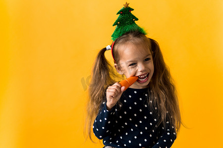 肖像积极开朗微笑快乐的小女学生圣诞树装饰圆点连衣裙咬着橙色背景上的橙色胡萝卜。