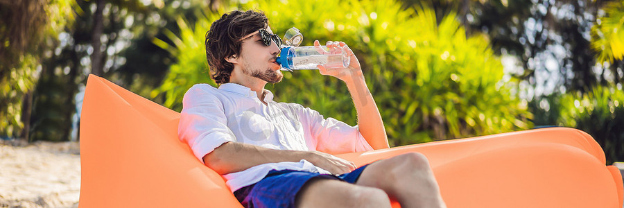 横幅，长格式夏季生活方式肖像，男人坐在热带岛屿海滩上的橙色充气沙发上喝水。