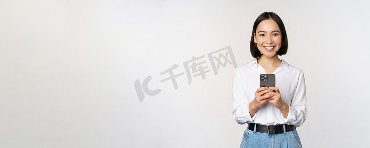 时尚的现代亚洲女孩使用手机应用程序，用手机聊天，微笑着，站在工作室背景下的白色衬衫