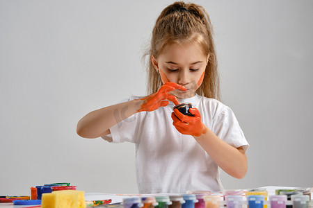 漫画美女涂色摄影照片_穿着白色 T 恤的小女孩坐在桌子旁，上面涂着什么和彩色颜料，正在给她的手涂色。