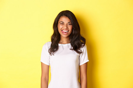 身穿白色T恤、无忧无虑的非洲裔美国女孩站在黄色背景上，露出舌头，微笑着，快乐地眨着眼睛