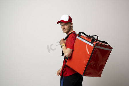 年轻的快递员穿着红色制服 T 恤，肩上挂着红色食品热袋，站在白色背景的工作室里。